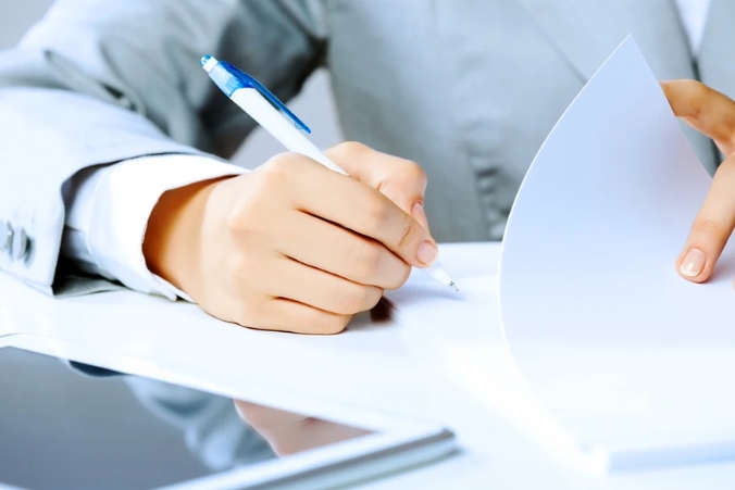 A person in a light suit writes on a sheet of paper at a desk, with a tablet and additional documents nearby.
