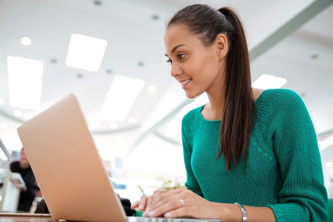 A young woman with long dark hair tied in a ponytail, wearing a green sweater, is smiling while working on a laptop in a bright indoor setting.