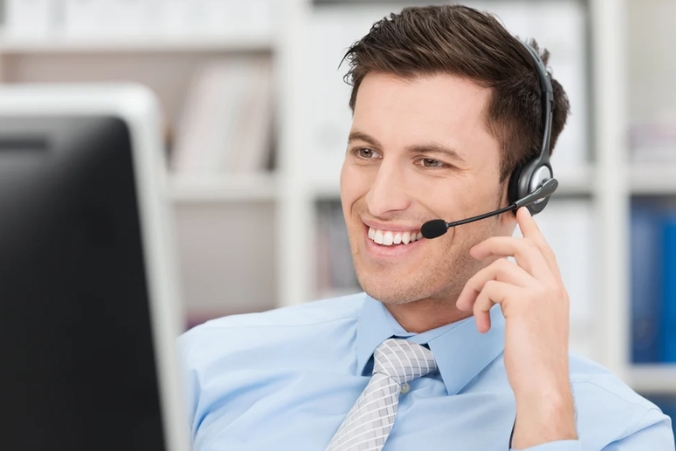 A smiling man wearing a headset and tie sits at a desk in front of a computer monitor, appearing to be on a call in an office setting.
