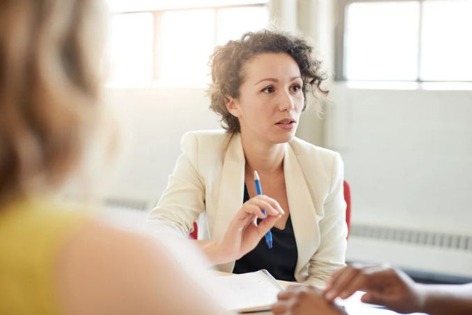 A woman with curly hair in a light blazer holds a pen and speaks during a meeting, seated at a table with two other people. Sunlight streams through large windows in the background.