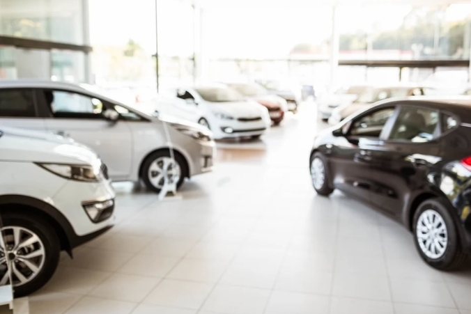 A blurred image of several cars displayed inside a bright, modern car showroom with white tiled floors and large glass windows.