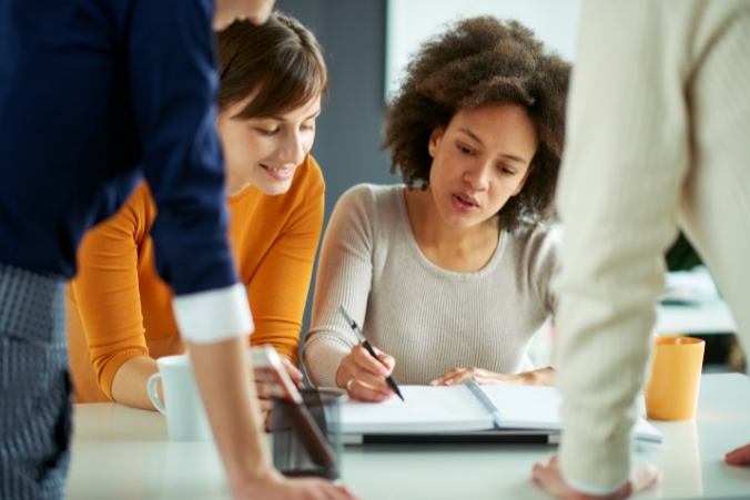 Four people are gathered around a table, collaborating. One woman is writing in a notebook while others watch and listen attentively. Cups and documents are on the table, suggesting a team meeting or group discussion.