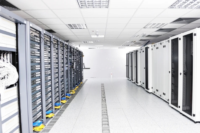 A brightly lit server room with rows of server racks on both sides, network cables neatly organized, and white tiled flooring with cable management channels in the center.